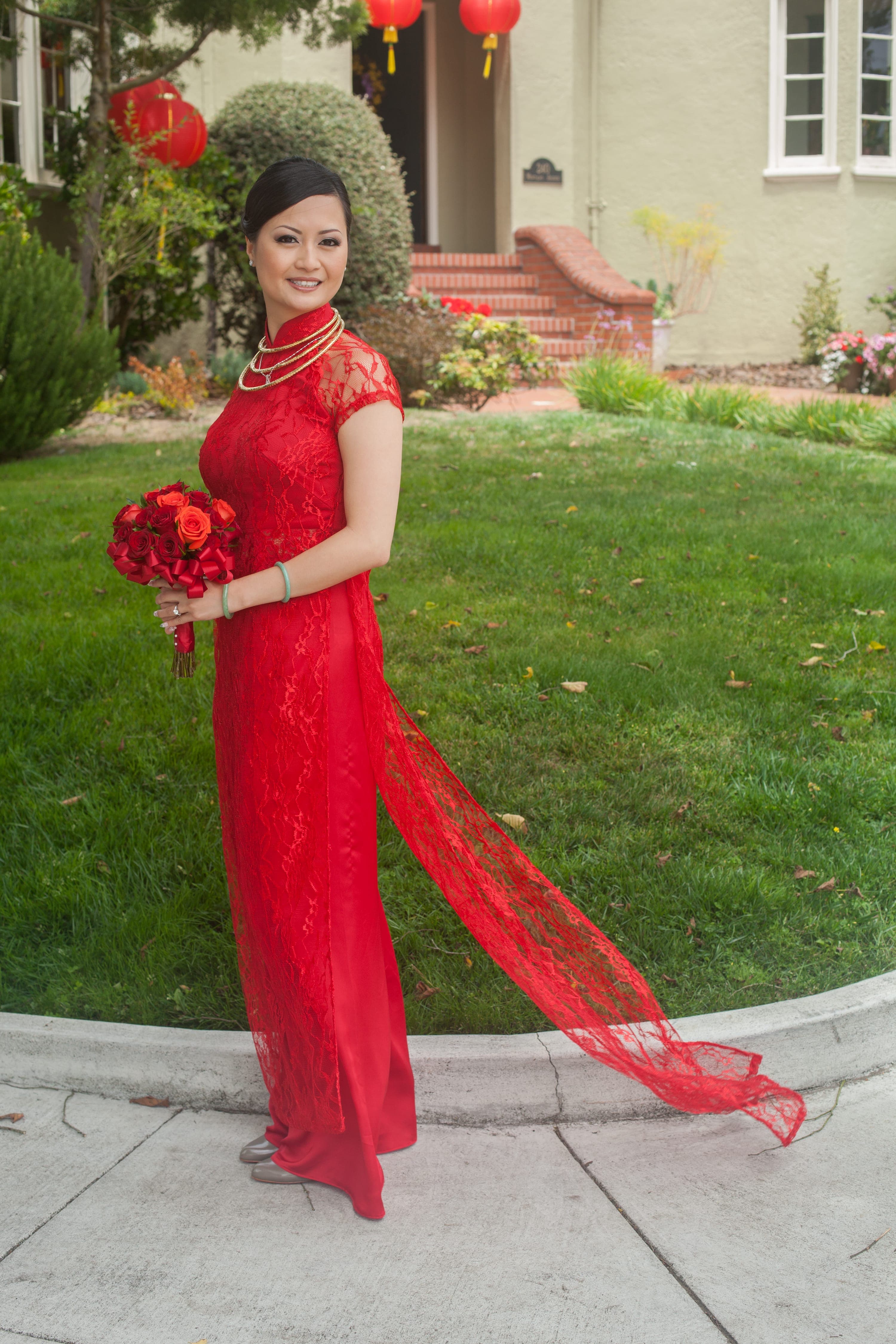 The bride standing on the sidewalk in a red lace áo dài holding a rose bouquet