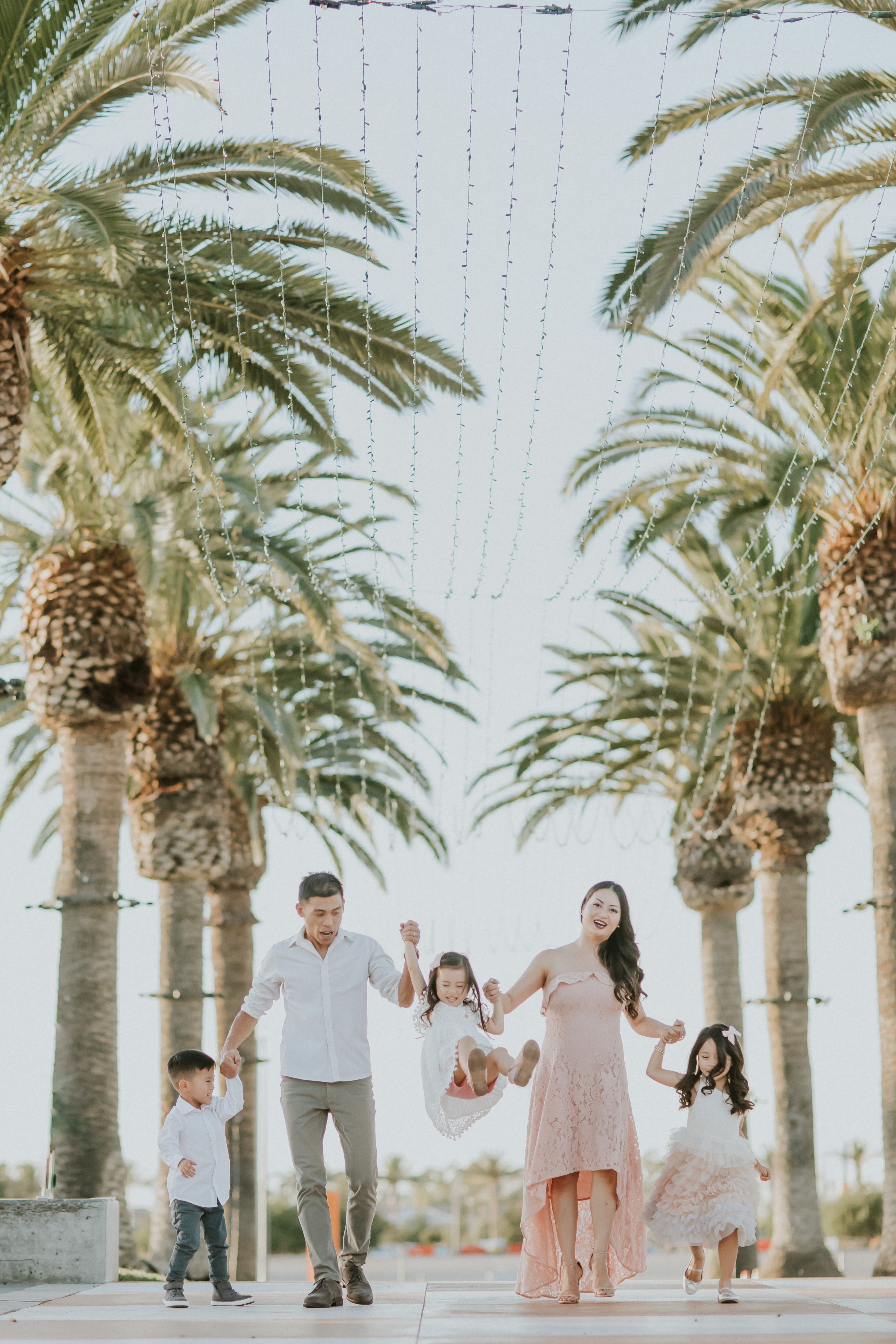 Parents swing their daughter between them under palm trees and string lights.