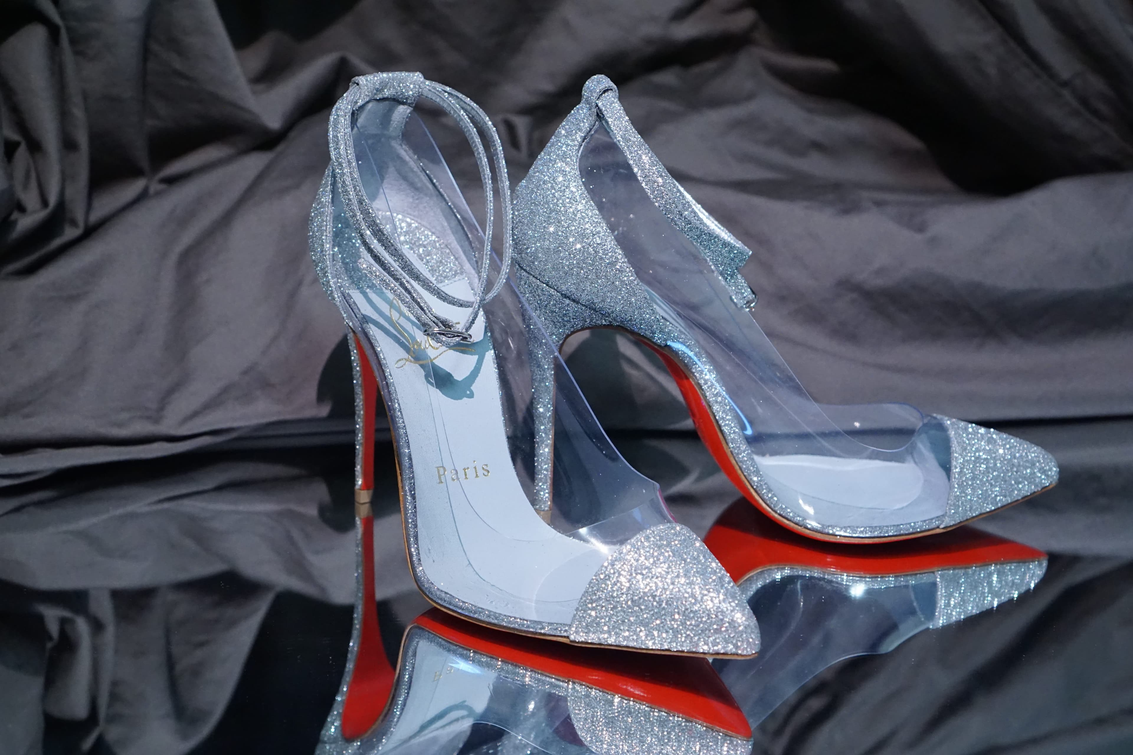 A pair of glitter Christian Louboutin pumps with clear PVC sides and ankle straps, reflected on a mirrored surface